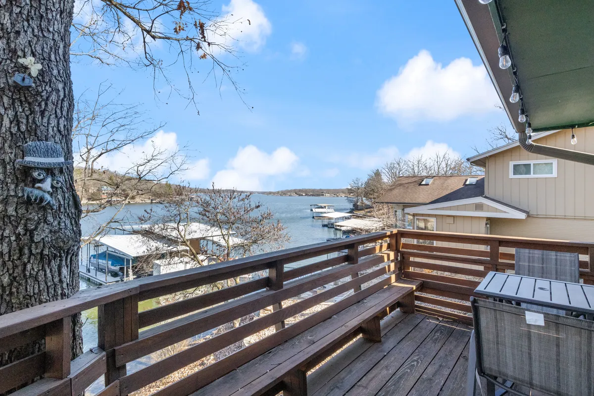 A tranquil lakefront scene captured from a rustic wooden deck, with a tree adorned in decorative ornaments in the foreground. The expansive view overlooks calm water bathed in the soft light of sunrise, inviting relaxation and early morning fishing directly from shore.