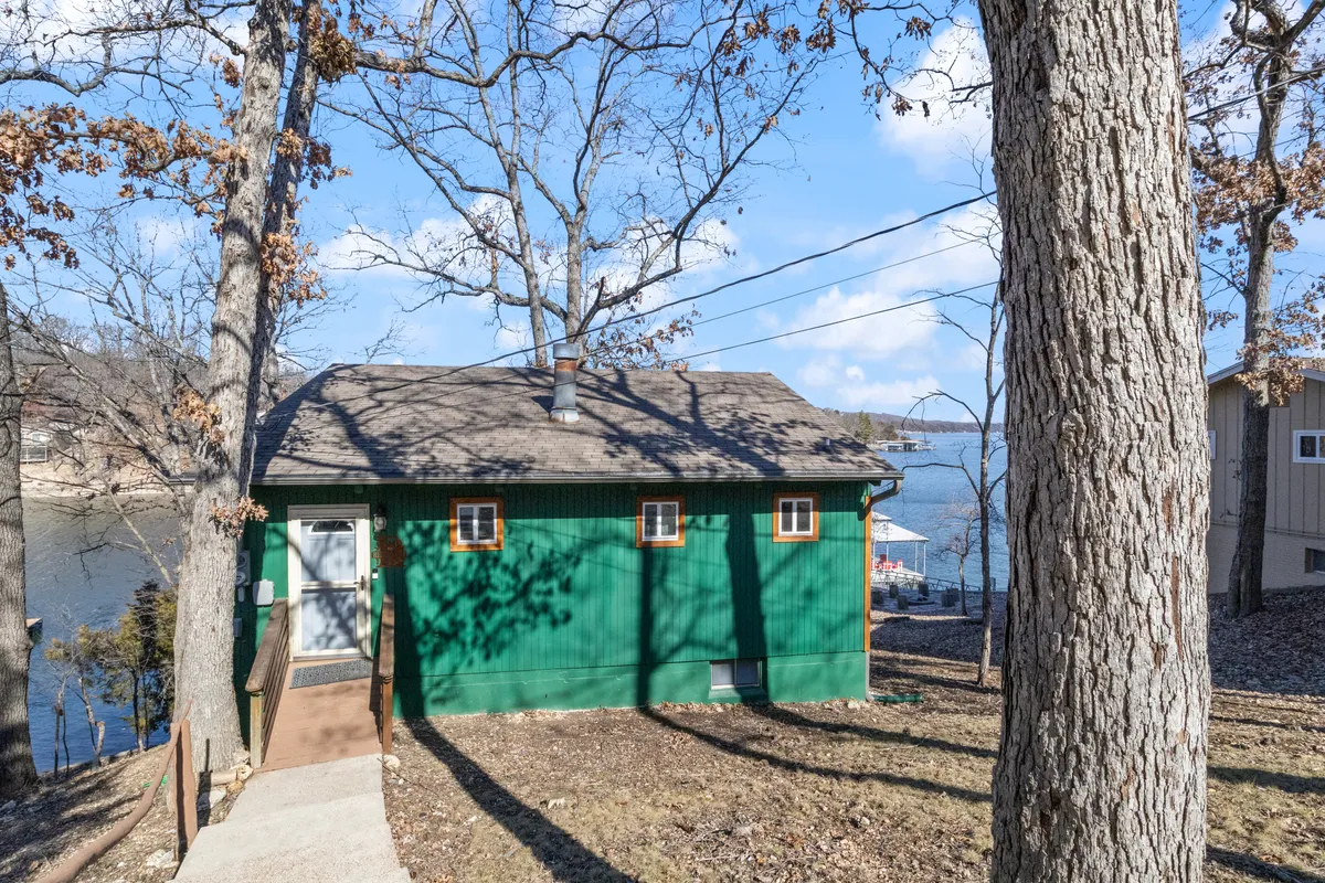 An exterior view of the green house nestled among mature trees, with a glimpse of the water in the background. The inviting setting emphasizes the rustic charm and immediate proximity to the lake, with subtle hints of outdoor living and tranquility.