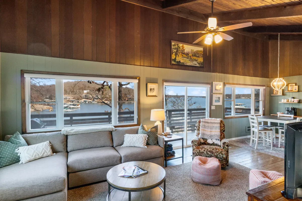A spacious living room featuring large windows that offer a sweeping view of the lake. The room is furnished with a comfortable sofa, artistic decor on the walls, and wood accents that create a warm, inviting atmosphere. Natural light floods the space, highlighting the rustic charm and encouraging gatherings and relaxation.