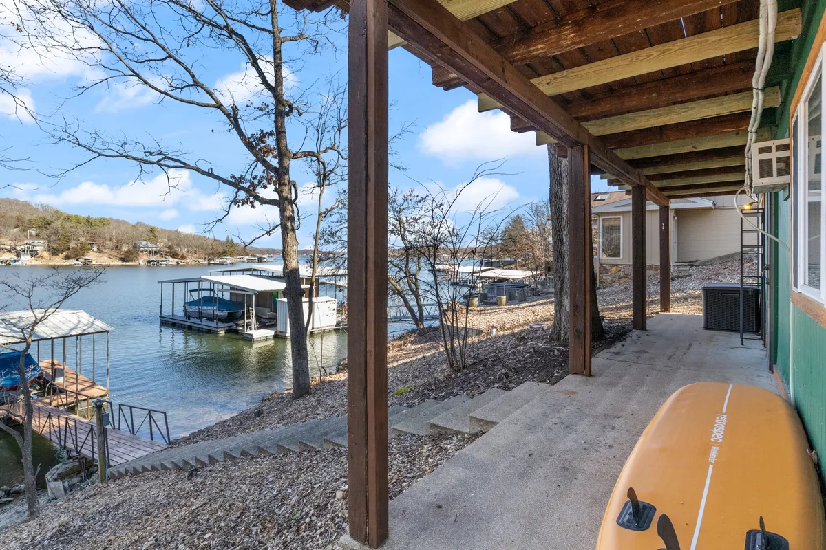 A picturesque lakeside setting with boathouses on the water’s edge, a staircase leading down to the shore, a kayak pulled up nearby, and trees framing the scene. The image conveys direct access to the water for fishing, boating, and a variety of lakeside activities.