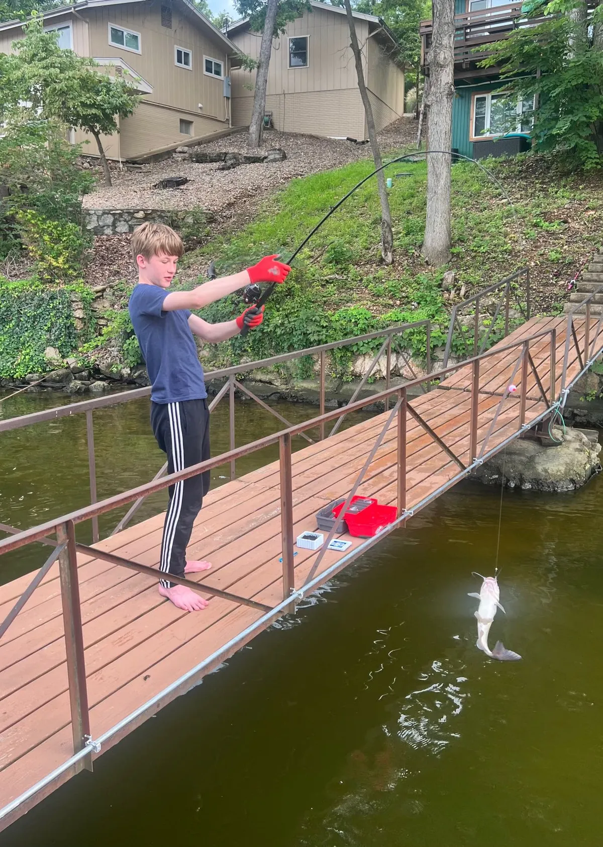 A joyful moment showing a young boy proudly holding a fishing rod with a freshly caught fish, standing near a footbridge by the water. The image exudes the excitement of fishing and outdoor play, highlighting intergenerational family fun.