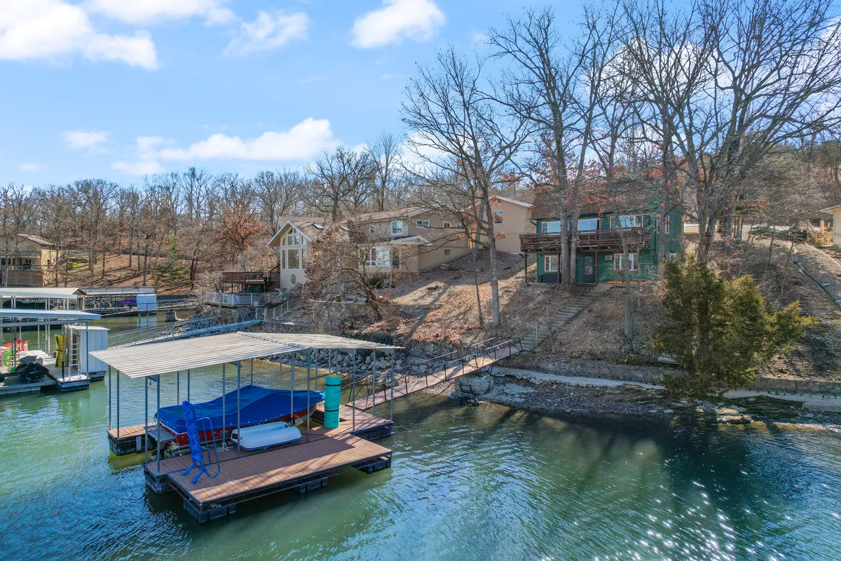 A serene view of the wooden dock stretching out over the lake, with hillside houses and lush trees in the background. The dock is clearly visible, emphasizing access to water activities and lounging in the sun.