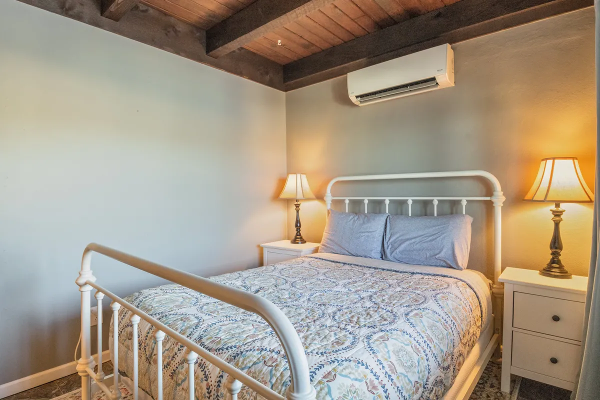 A tranquil bedroom with a bed adorned with decorative bedding, warm wooden beams on the ceiling, and nightstands with soft lamps. Gentle natural light fills the space, creating an atmosphere of calm and relaxation ideal for couples or solo travelers seeking peaceful rest.