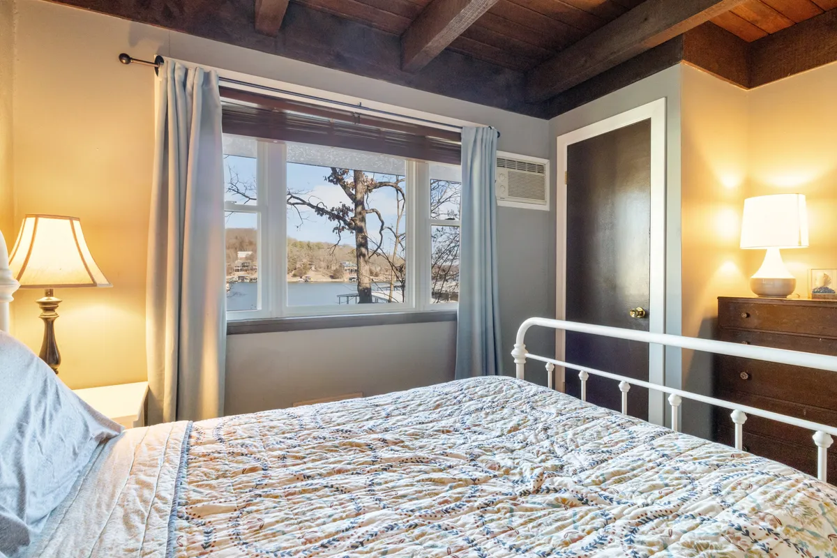A cozy bedroom featuring a bed with a patterned quilt, a window that frames a tranquil outdoor view, and a softly glowing lamp on the nightstand. The rustic wooden elements and serene natural light create a calming atmosphere ideal for unwinding after a day outdoors.