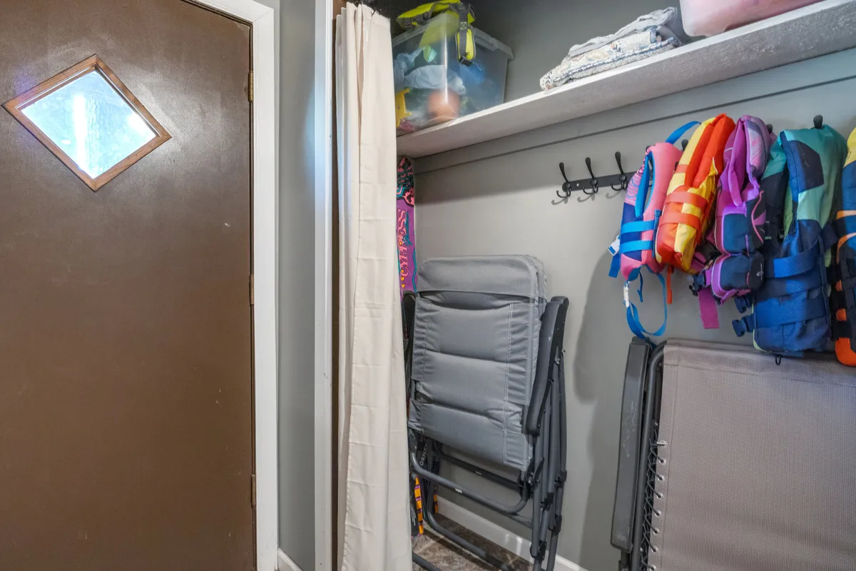 A practical entryway or mudroom scene with a door featuring a diamond window, colorful life jackets, folding chairs, and shelves for storage. The space is ready for guests to prepare for lake adventures with gear at hand.