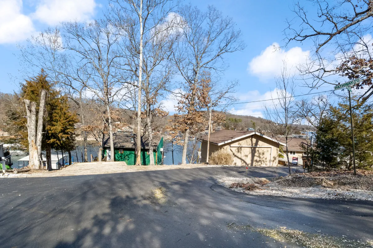 A sweeping view across the lake framed by a tree line, with adjoining properties and a quiet roadway in the background. The tranquil atmosphere suggests leisurely mornings, fishing, or simply soaking in the scenery from a prime waterfront position.